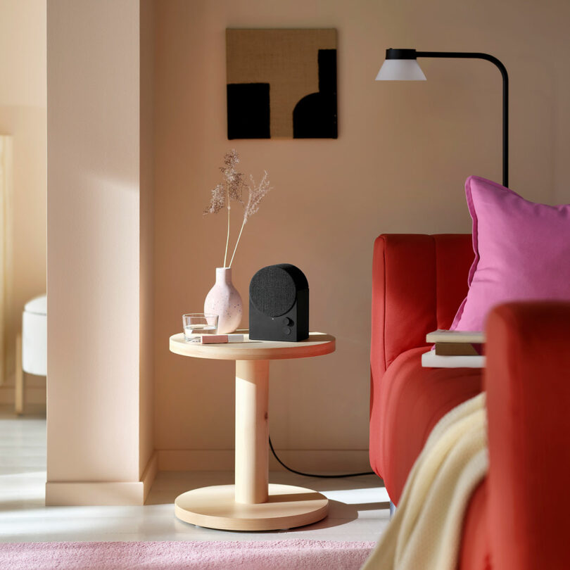 A round wooden side table with a pink vase, dried flowers, a glass of water, a black speaker, and books beside a red sofa with pink cushions in a modern living room