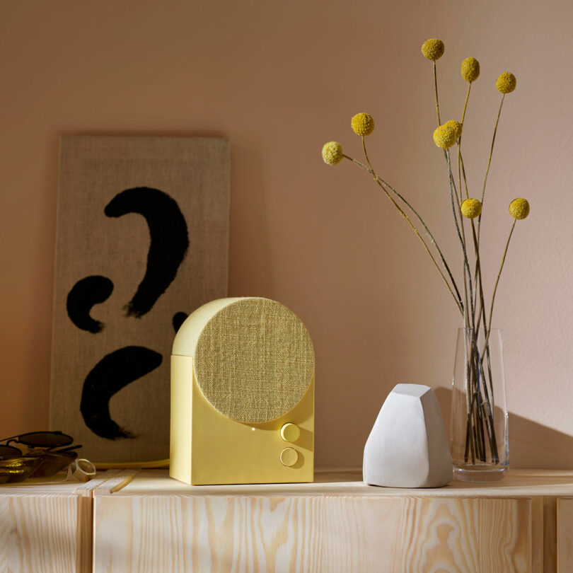 A yellow speaker sits on a wooden shelf next to a minimalist stone sculpture, a glass vase with yellow billy button flowers, and abstract black-and-white artwork