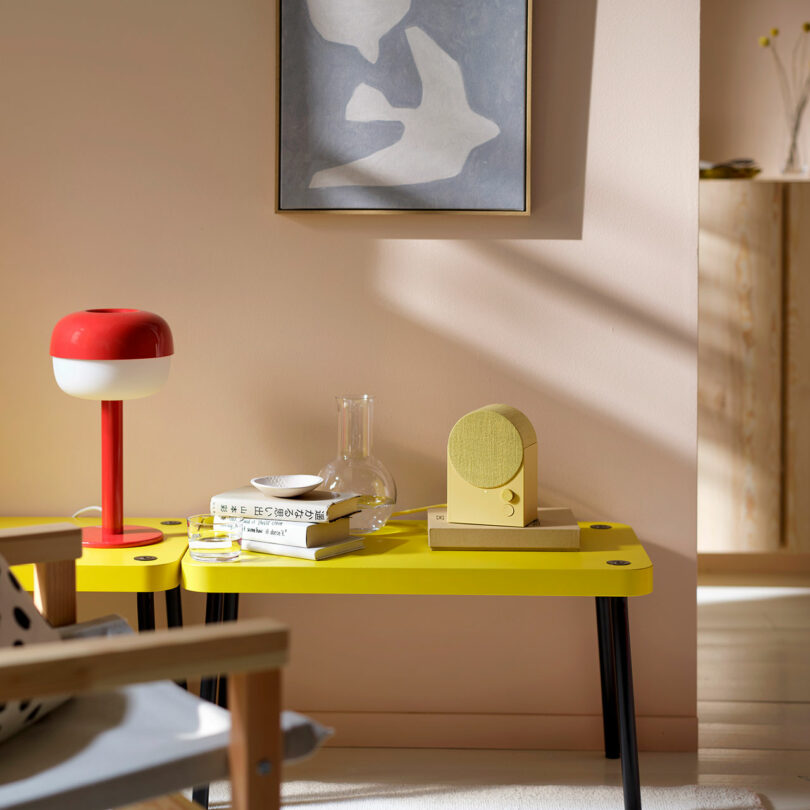 A yellow table with a red and white lamp, books, a glass carafe, and a yellow speaker, set against a pale wall with abstract art and soft natural light