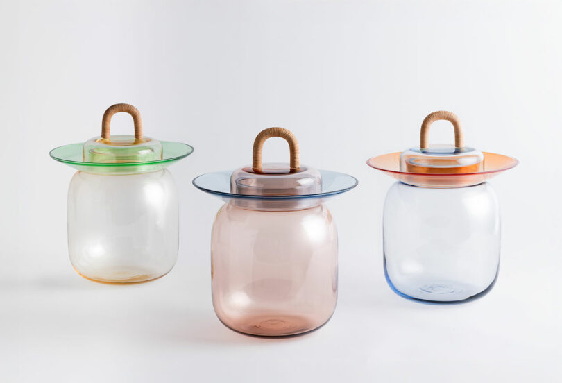 Three translucent glass containers with wide, flat lids in green, blue, and orange, each featuring a rope handle, are displayed against a white background.