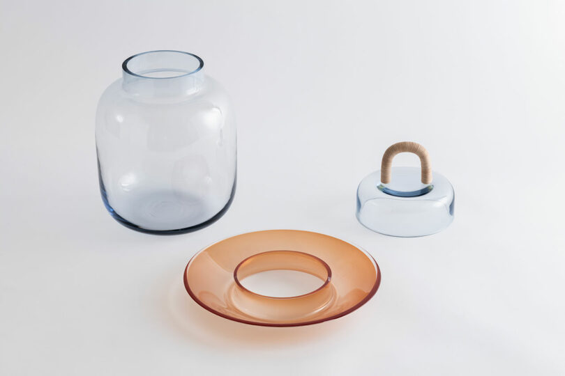 Three modern glass objects are displayed on a white surface: a blue vase, an orange circular plate with a hollow center, and a blue dome with a handle wrapped in cord.