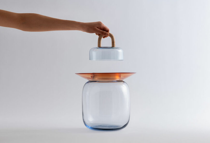 A hand lifts a glass dome with a rope handle from the top of a transparent glass container with a wide orange rim, set against a plain white background.
