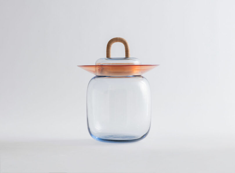 Clear glass jar with rounded edges, an orange circular lid, and a looped handle wrapped in natural fiber, set against a plain white background.
