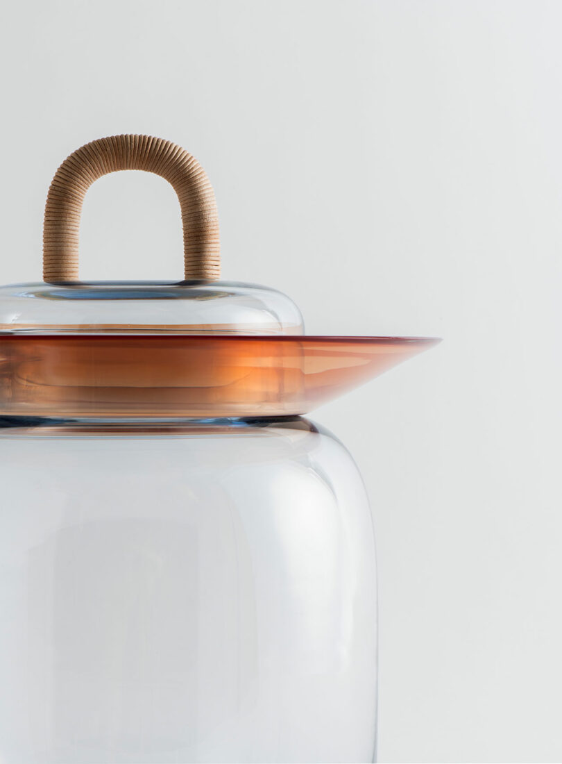 A clear glass jar with a translucent orange lid and a handle wrapped in light brown cord, set against a plain white background.