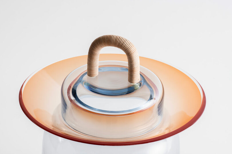 A glass container with an orange-edged lid and a central handle wrapped in beige cord, set against a plain white background.