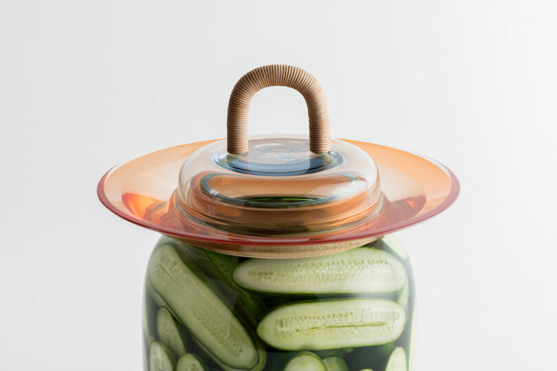 A glass jar filled with cucumbers is topped with an orange and clear lid featuring a brown handle.
