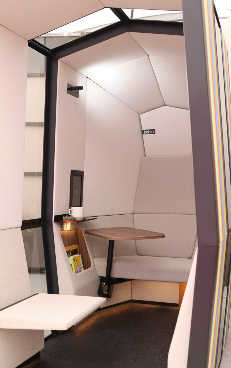 The Kabin is a modern, compact work pod featuring a cushioned bench, wooden table, wall-mounted screen, reading light, and magazine holder—perfectly designed for individual use.