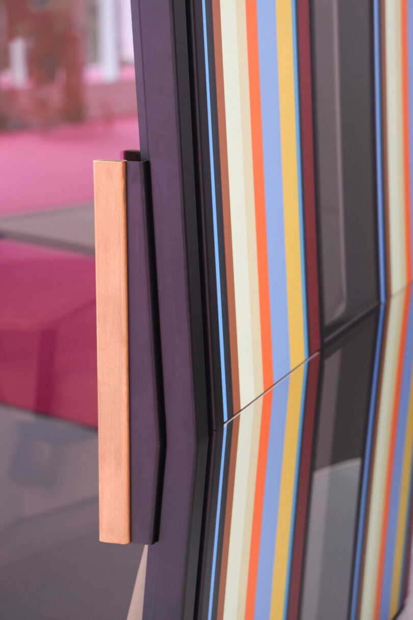 Close-up of a Kabin cabinet door with a wooden handle, featuring a surface decorated with vertical pastel and bold colored stripes and its reflection below.