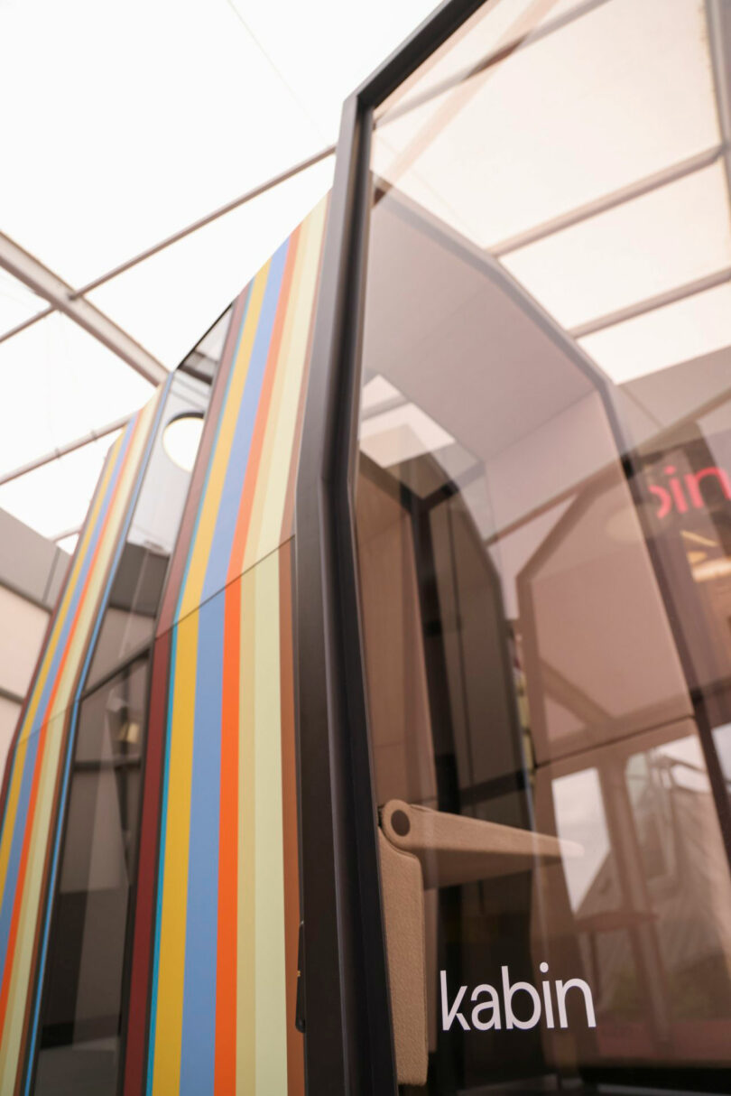 A modern Kabin phone booth with a glass door and vertical colorful stripes, labeled "kabin," set inside a bright indoor space.