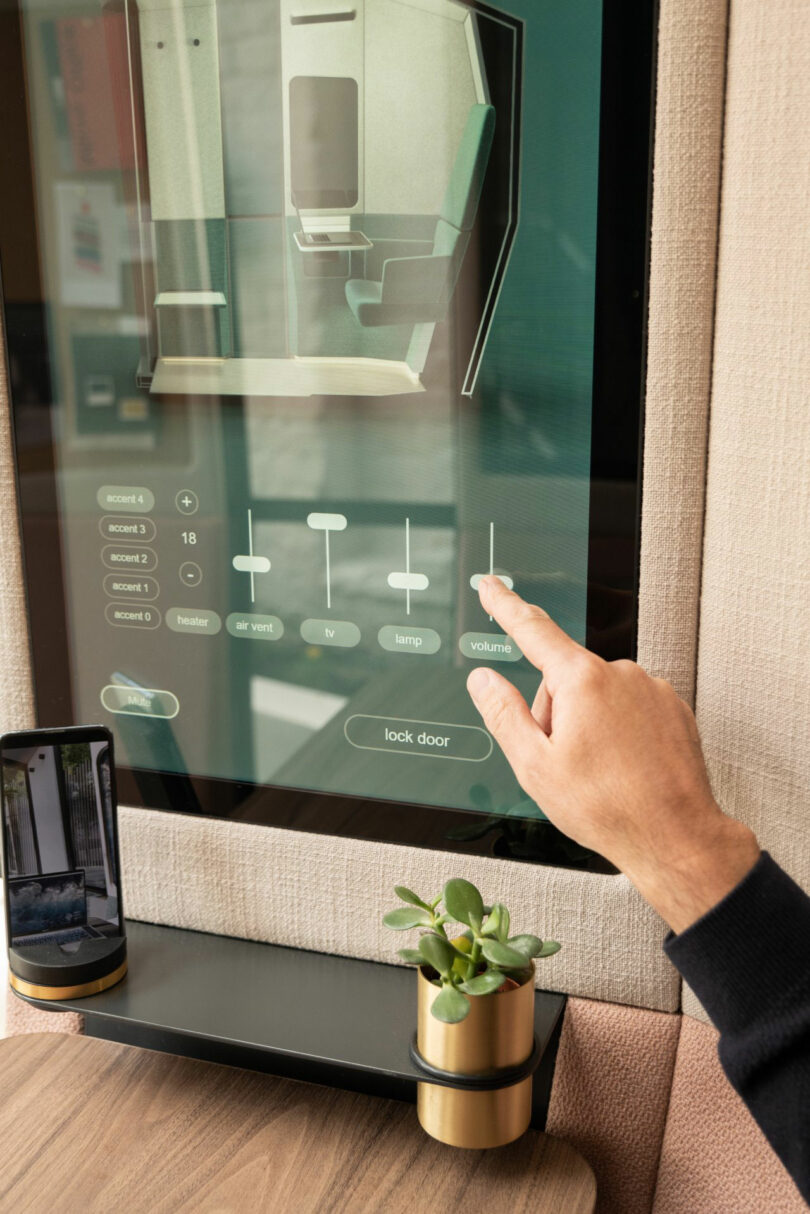 A person adjusts the volume slider on a Kabin wall-mounted touchscreen control panel, with a smartphone and a small potted plant resting on a nearby shelf.