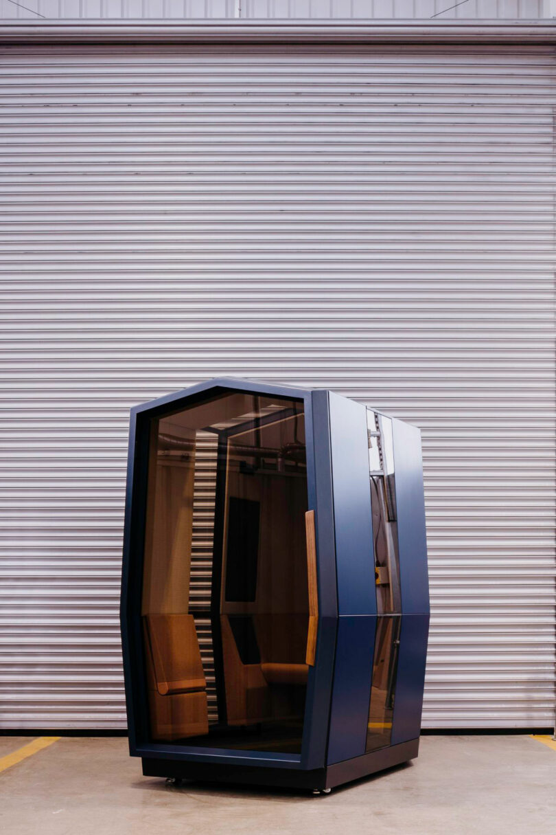 A modern Kabin, featuring glass walls and a wooden interior, stands on a concrete floor in front of a large metal roll-up door.