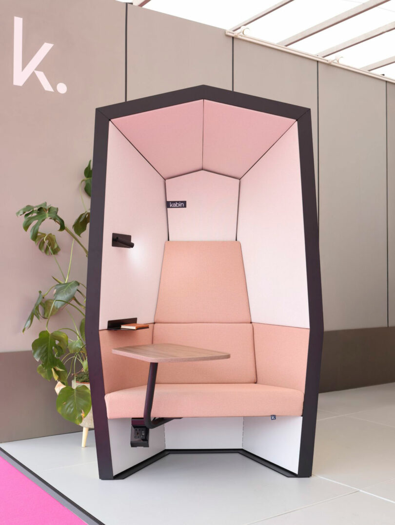 A modern, pink upholstered Kabin privacy pod with a built-in table and outlets sits on a gray floor next to a green potted plant.