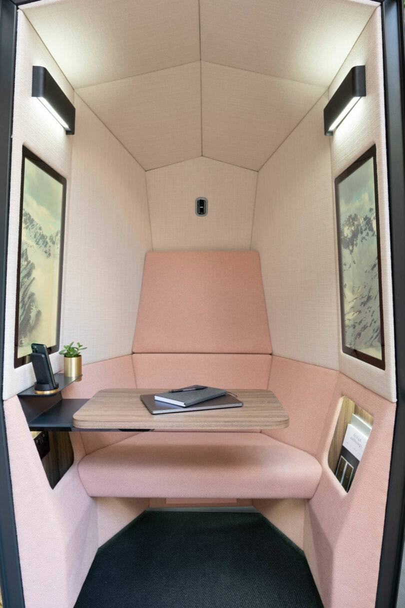 A small, private Kabin office booth with a cushioned seat, desk, notebook, phone, plant, and nature-themed wall art under built-in lights.