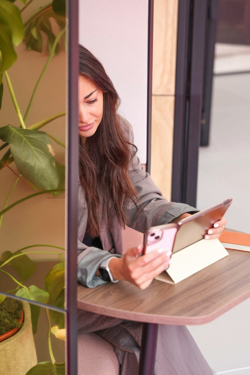A woman sits at a small Kabin table with a tablet in a stand, holding a smartphone, next to a green leafy plant.