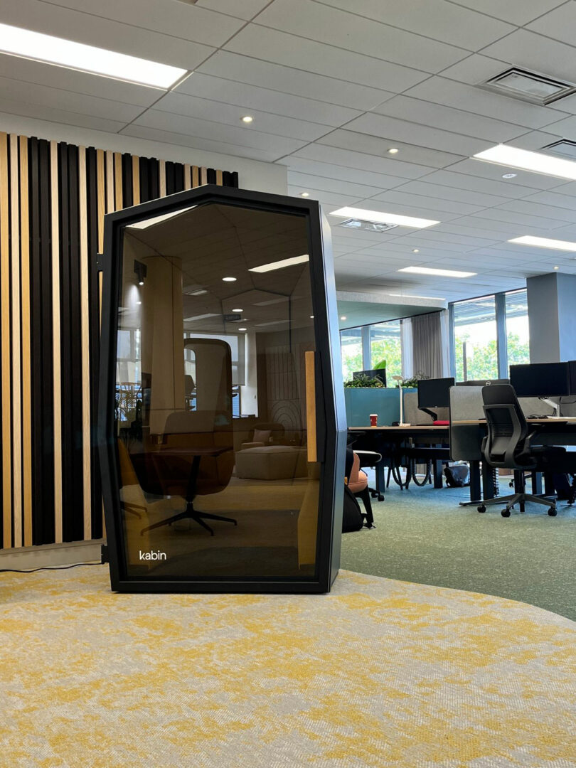 A modern office space featuring a black, glass-fronted Kabin phone booth, surrounded by desks, chairs, and large windows.