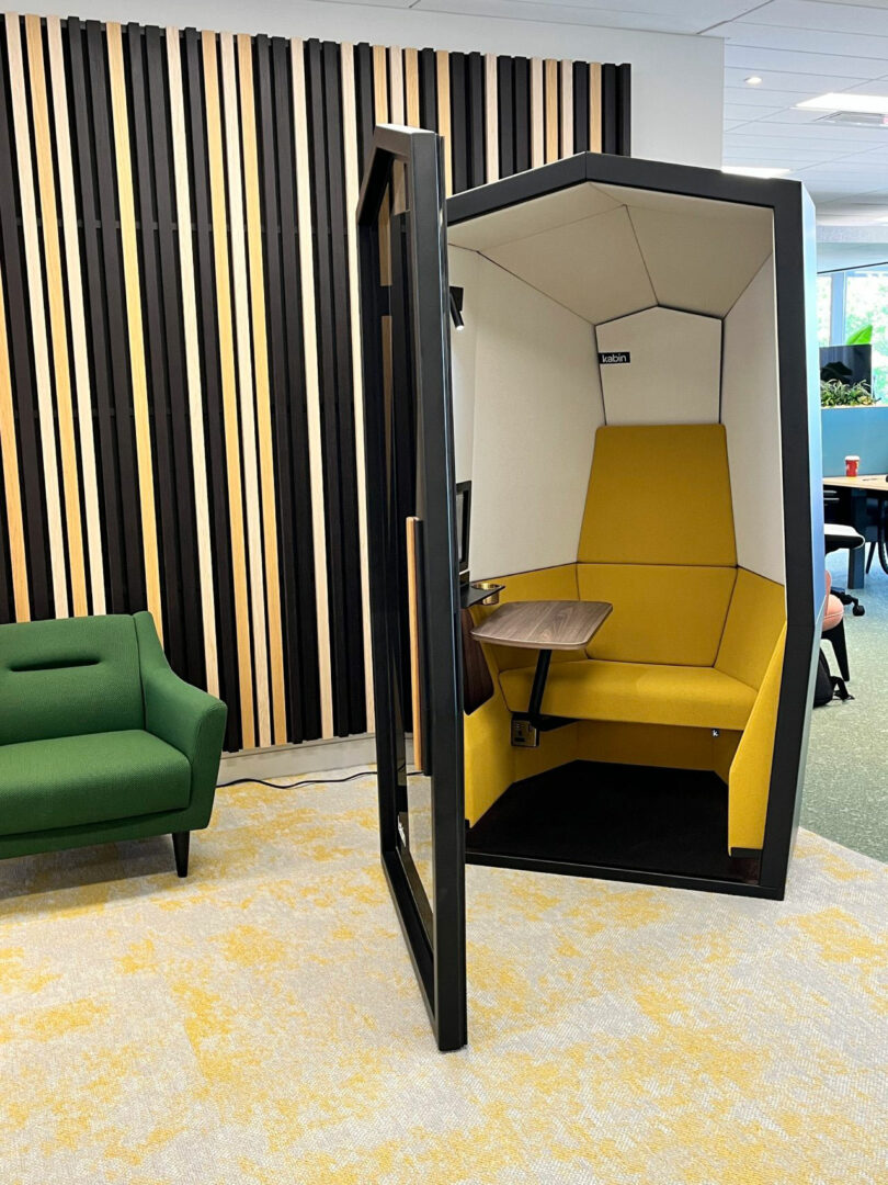 A modern office setting with a green sofa and a yellow Kabin privacy pod featuring a small desk and seating, positioned on a carpeted floor near a decorative wooden wall.