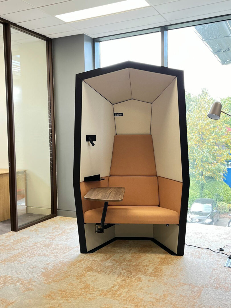 A modern Kabin office pod with beige upholstery, a wood desk, and partial enclosure sits near large windows overlooking trees and a parking lot.