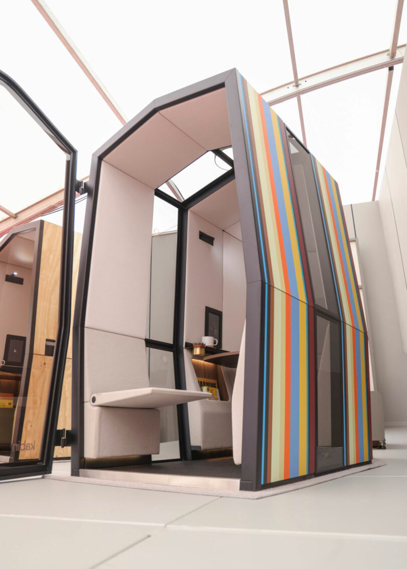 A modern Kabin office pod with glass walls and colorful vertical stripes on the exterior, featuring a built-in bench and desk inside.