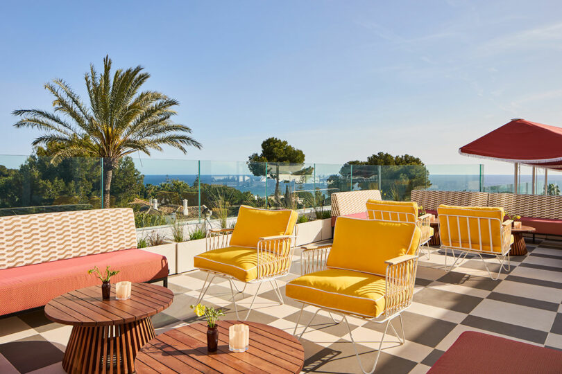 Outdoor terrace at Kimpton Los Monteros with yellow lounge chairs, patterned sofas, wooden tables, and potted plants overlooking the sea under a clear sky, with palm trees in the background.