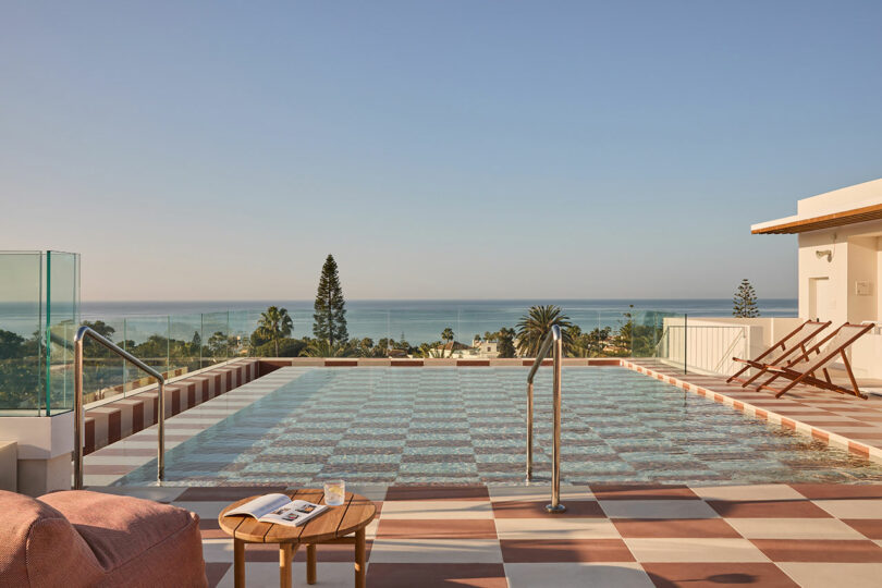 Rooftop pool with checkered tile deck, lounge chairs, and a table at Kimpton Los Monteros, overlooking the ocean and palm trees under a clear sky.