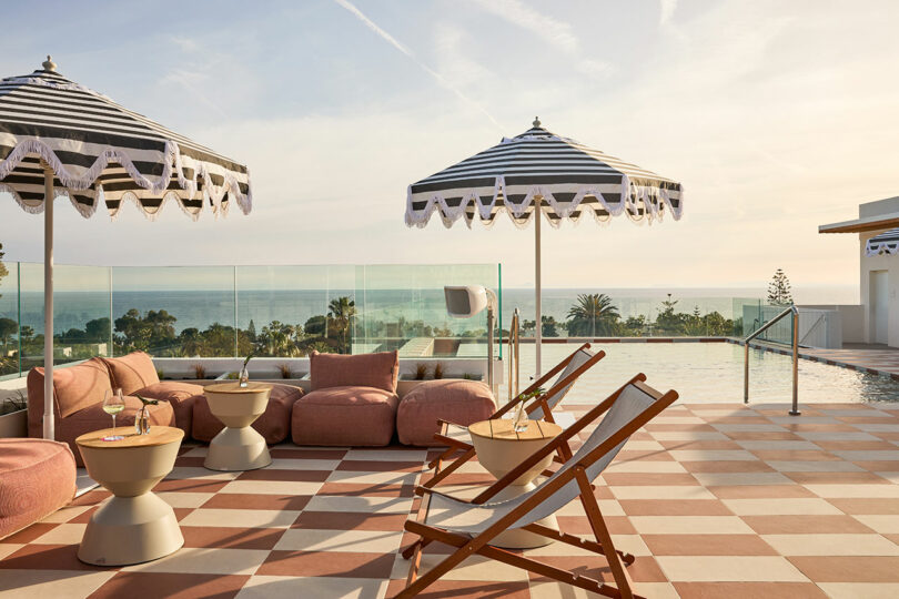 Outdoor lounge area at Kimpton Los Monteros featuring striped umbrellas, cushioned seating, deck chairs, and small tables overlooking a pool and ocean view under a clear sky.