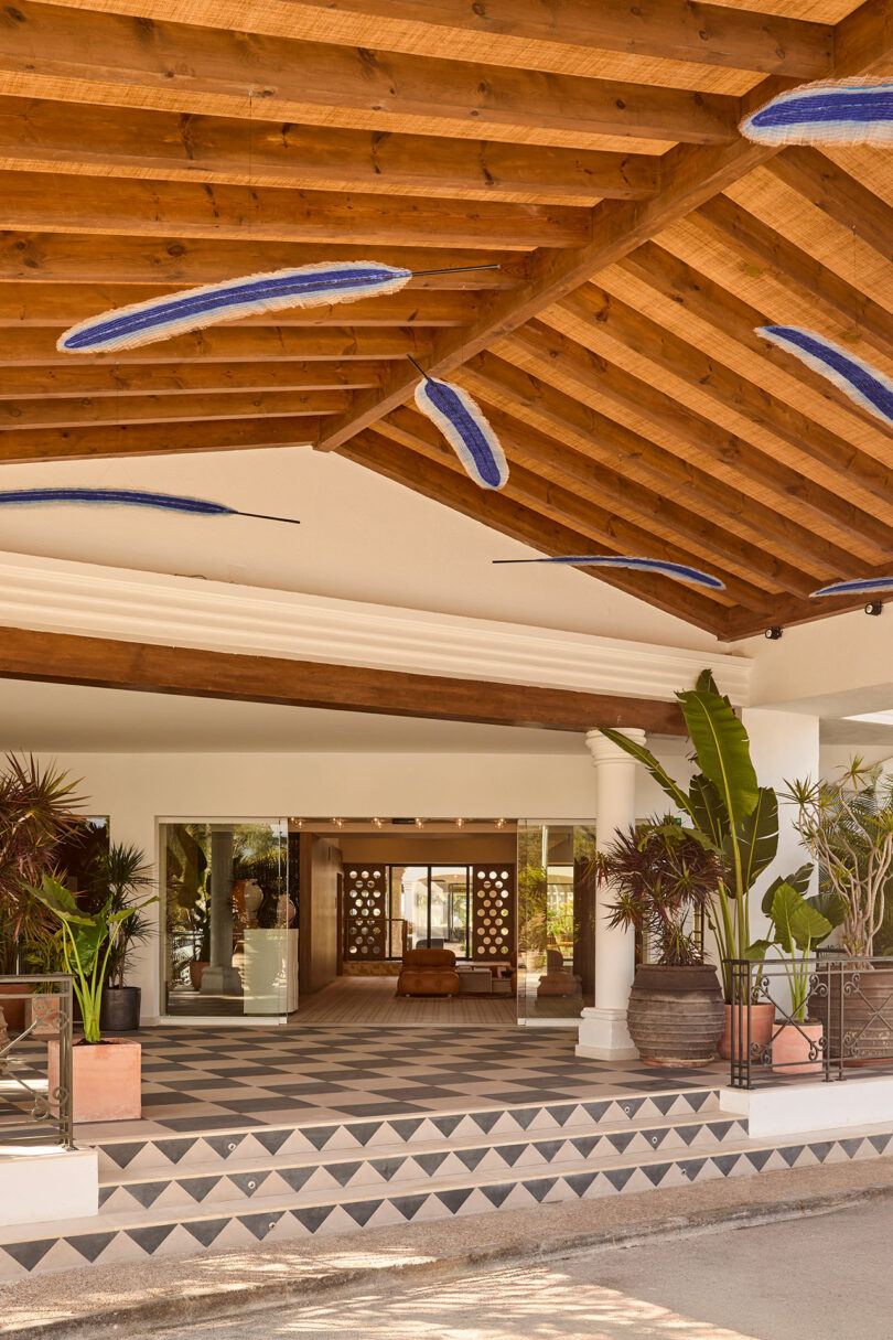 Covered entrance to Kimpton Los Monteros, featuring wooden beams, potted plants, geometric tile steps, and glass doors leading into a stylish lobby.
