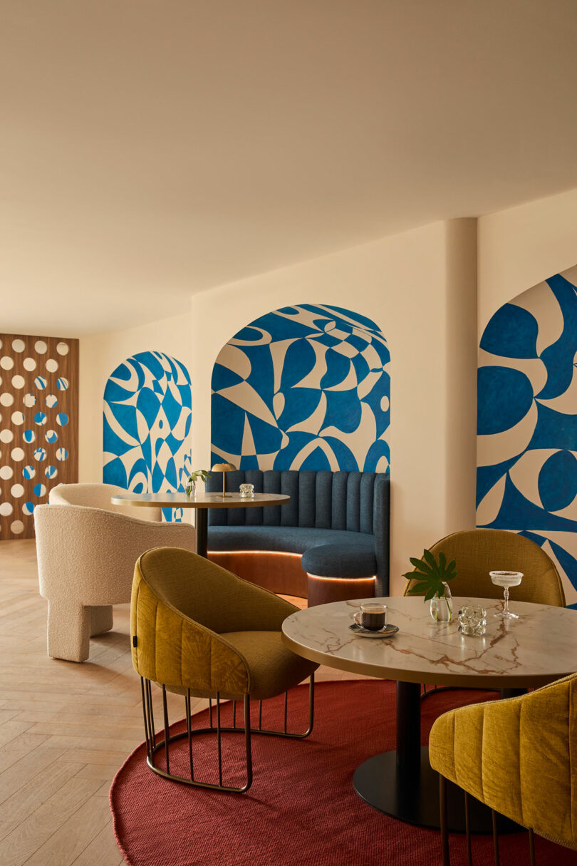 Modern cafe interior at Kimpton Los Monteros with round tables, yellow and white chairs, a red rug, and blue abstract wall art set within arched alcoves.