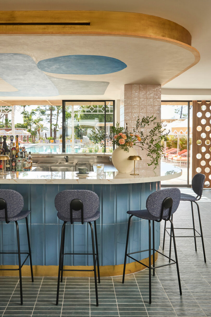 Modern bar area at Kimpton Los Monteros with a curved blue-tiled counter, four bar stools, a large floral arrangement, and floor-to-ceiling windows overlooking an outdoor pool.