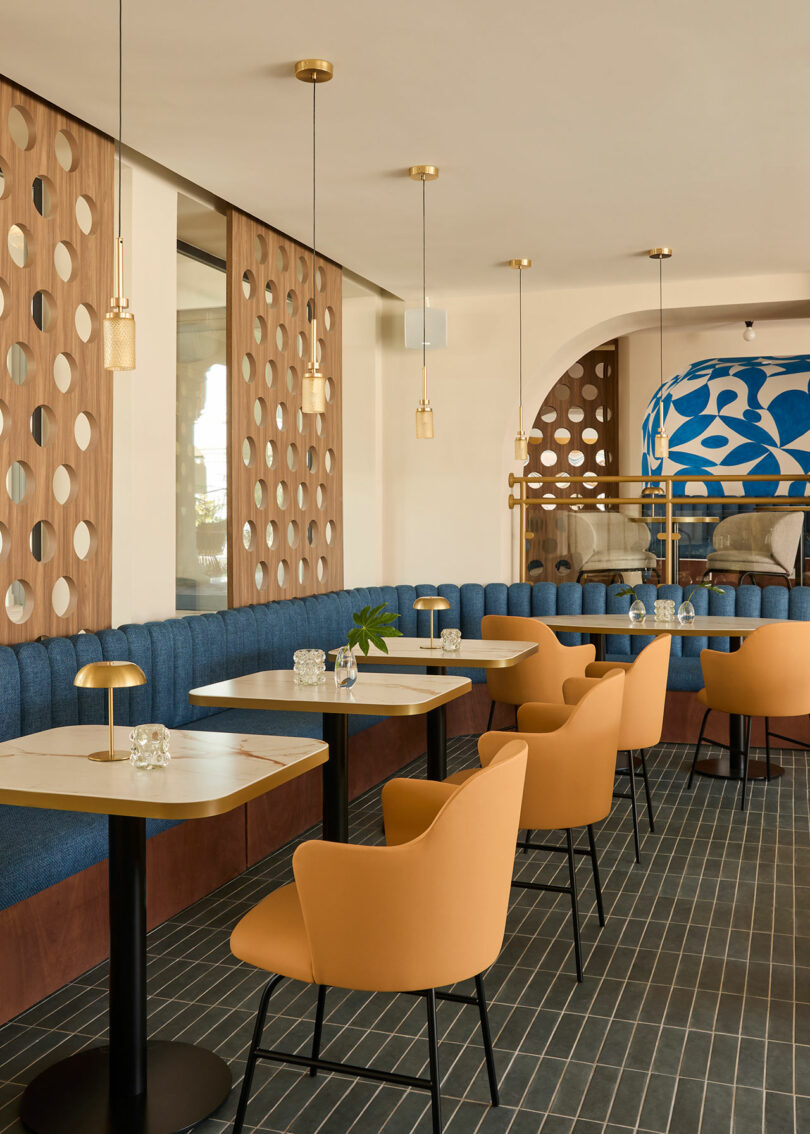Modern café interior inspired by Kimpton Los Monteros, featuring blue upholstered bench seating, mustard yellow chairs, pendant lights, wooden dividers with circular cutouts, and a blue and white mural in the background.