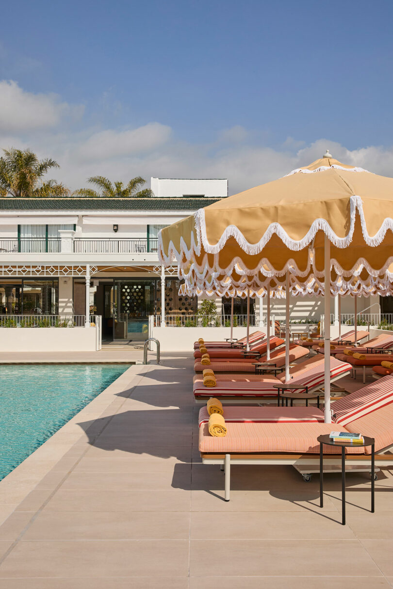 Poolside loungers with pink cushions and rolled yellow towels are arranged under a large fringed yellow umbrella beside a clear swimming pool at Kimpton Los Monteros resort.