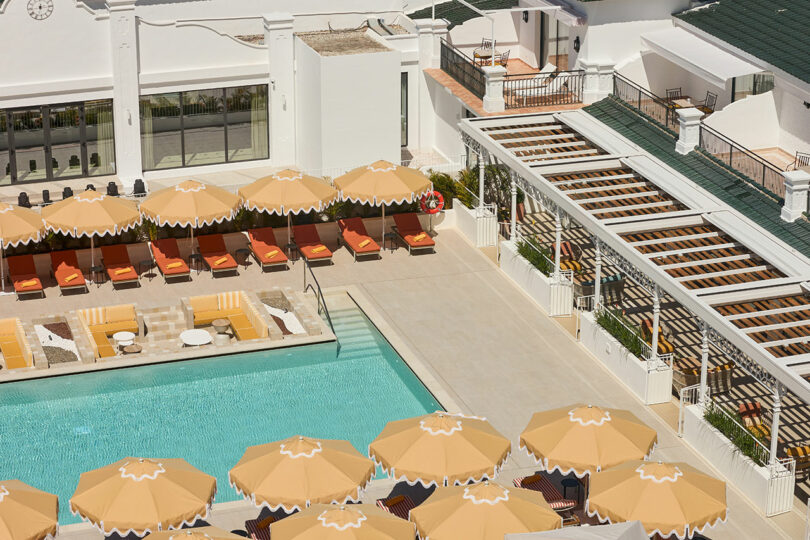 Aerial view of Kimpton Los Monteros poolside area with orange lounge chairs and sun umbrellas, surrounded by white buildings and a covered patio.
