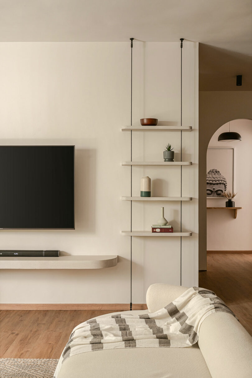 A modern living room with a wall-mounted TV, floating shelves holding decorative items, a white sofa with a patterned throw, and an arched doorway leading to another room.