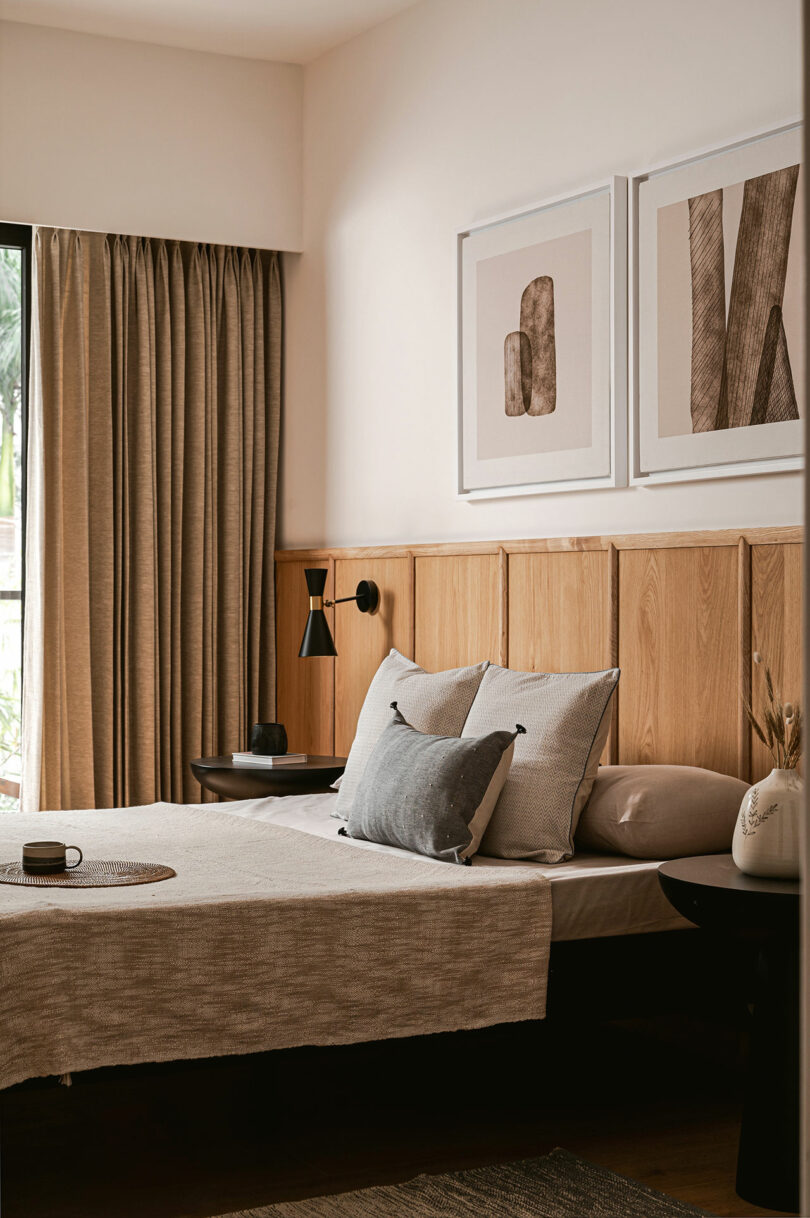 A modern bedroom with a wooden headboard, neutral bedding, two framed abstract artworks on the wall, and beige curtains covering a window.