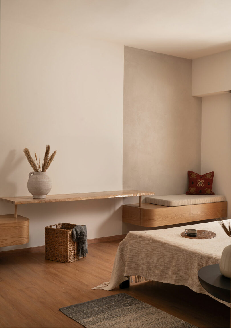 Minimalist bedroom with a floating wooden desk, a bench with a red cushion, woven basket, neutral linens, and decorative vase with dried grass on the desk.
