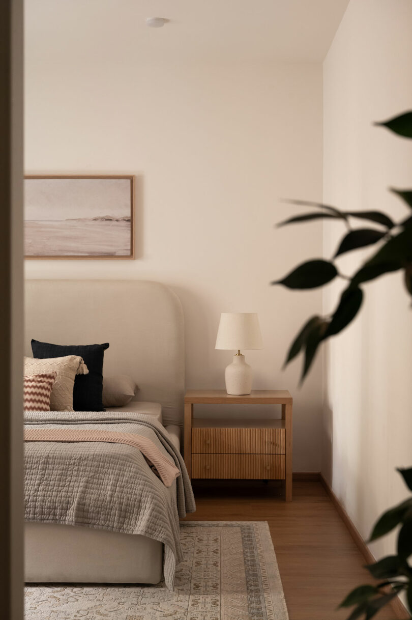 A neatly made bed with neutral bedding, a wooden nightstand with a lamp, a framed picture on the wall, and a plant partially visible in the foreground.