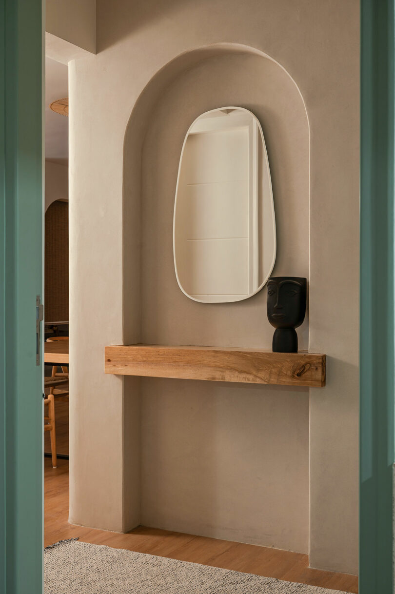 A modern entryway with a curved niche, wooden shelf, abstract black vase, and irregular oval mirror above light wood flooring.