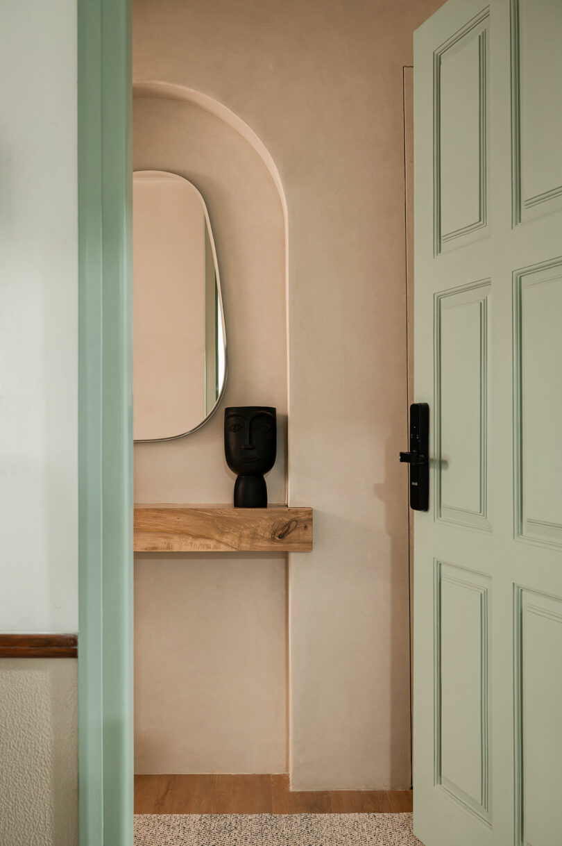 A mint green door opens to a niche with a wooden shelf, a sculptural black vase, and an arched mirror on a beige wall.