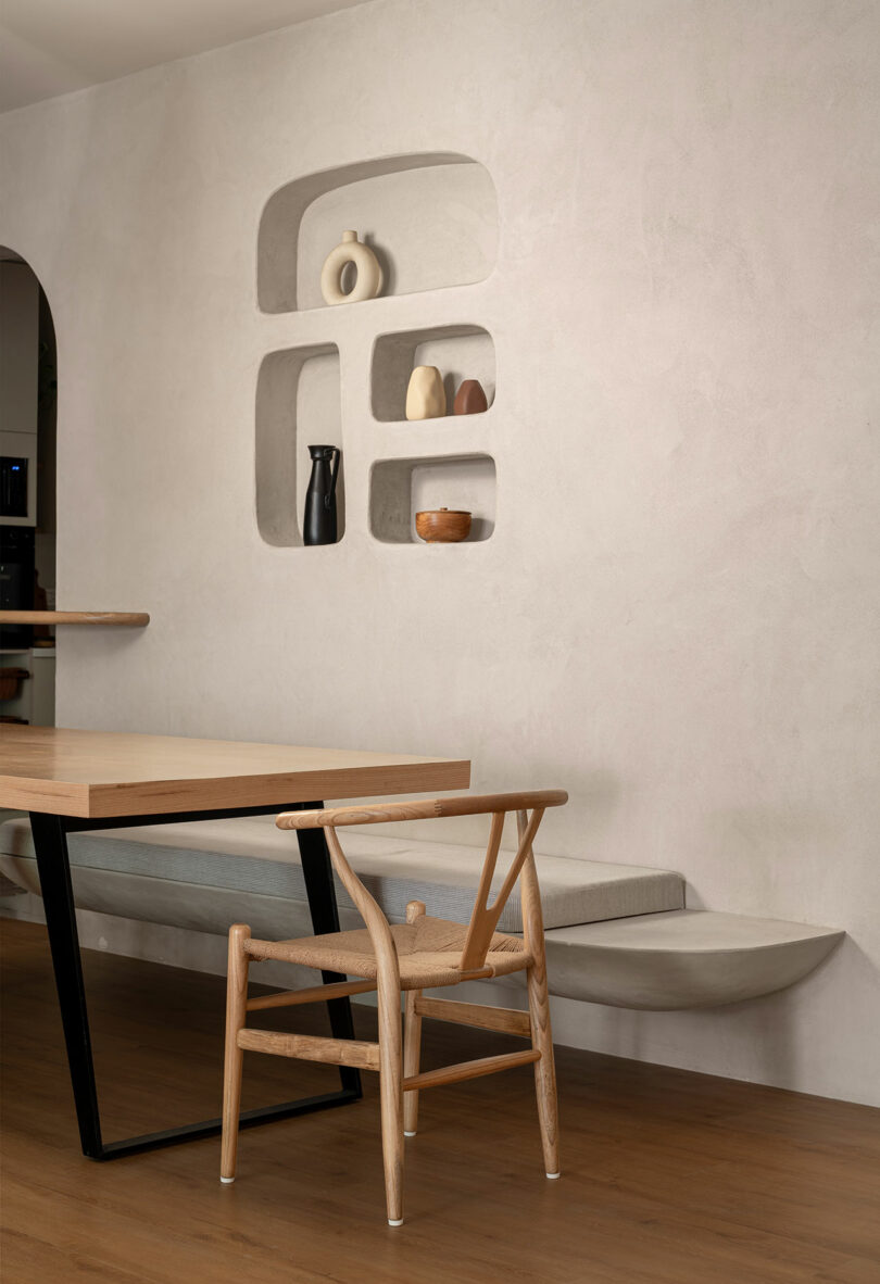 Minimalist dining table with a wooden table, wooden chair, built-in wall shelves showing simple ceramic and floating concrete bench against light textured wall.