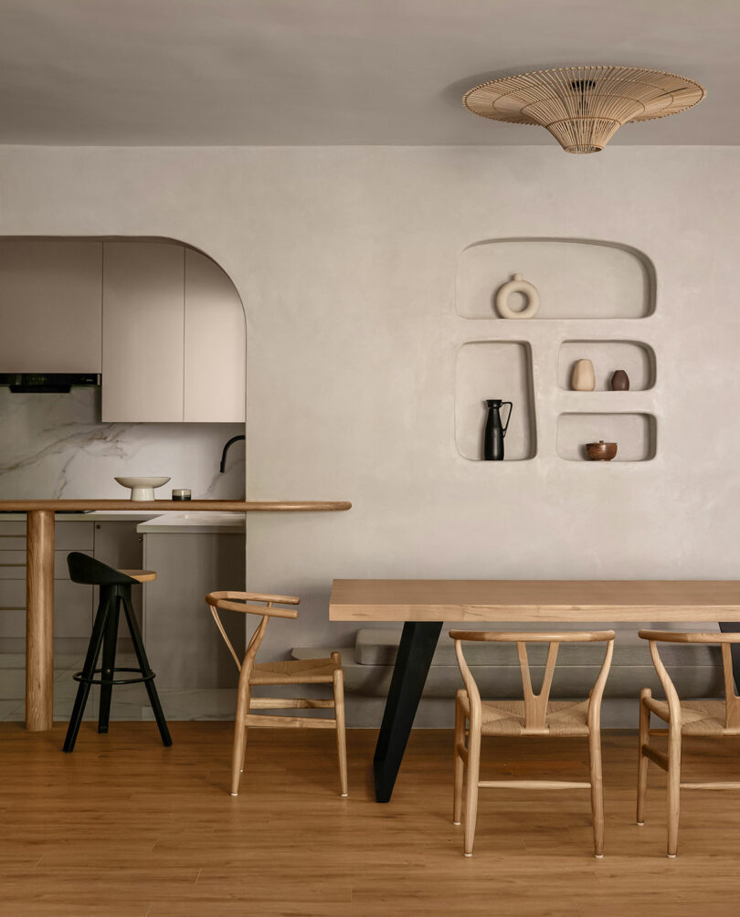 Minimalist dining area with light wood furniture, built-in wall shelves displaying vases, and a view into a small kitchen with neutral tones and a wooden breakfast bar.