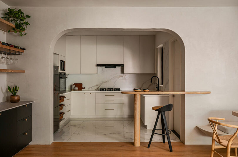 Modern kitchen with white cabinets, marble floor, built-in appliances, a black stool at a wooden breakfast bar, and a potted plant on the counter.