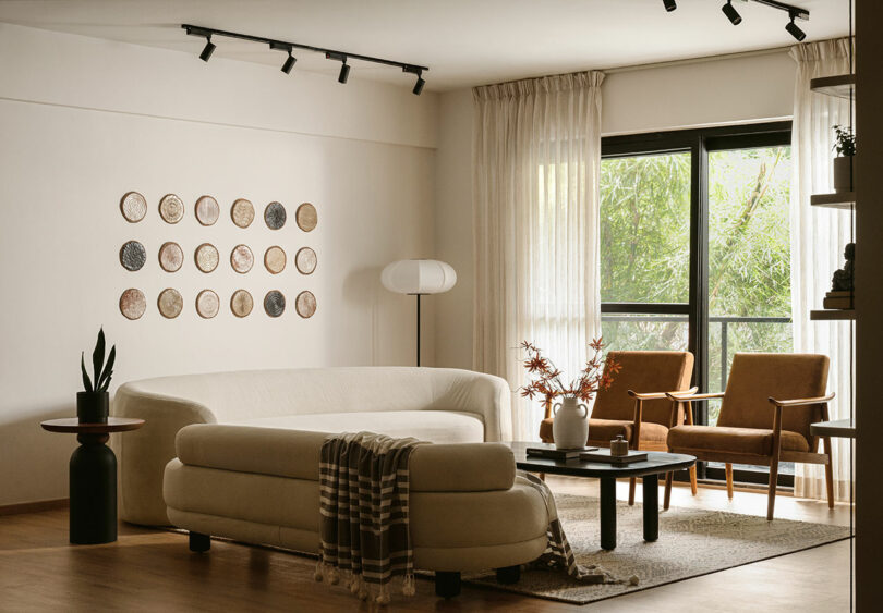 A modern living room with a white curved sofa, round coffee table, two brown armchairs, a floor lamp, wall art, and large window with sheer curtains letting in natural light.