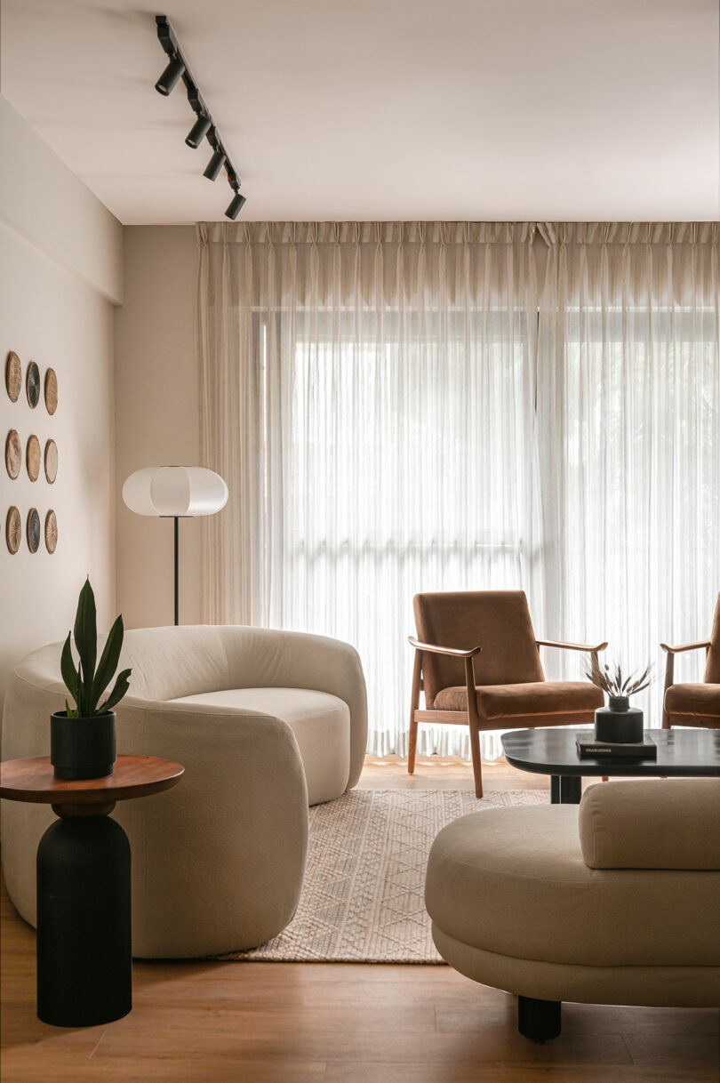 Modern living room with beige curved sofas, a round black table with a potted plant, two brown chairs, and sheer curtains covering large windows.