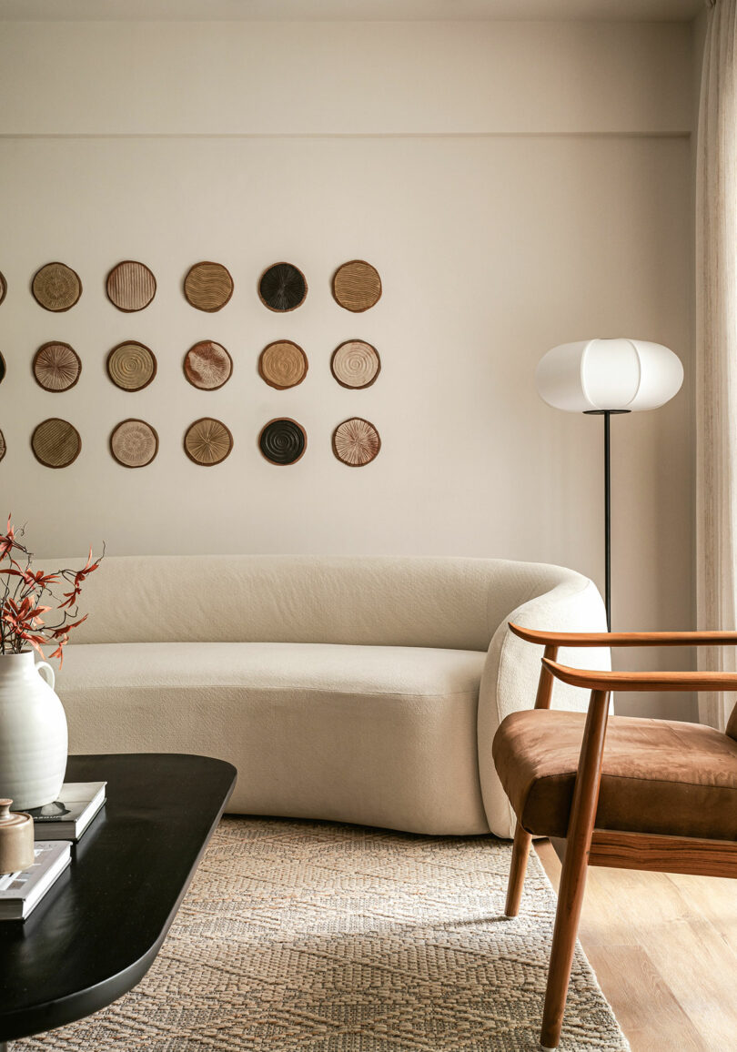 A modern living room with a curved beige sofa, a wooden chair, a black coffee table, a floor lamp, and decorative round wall art above the sofa.
