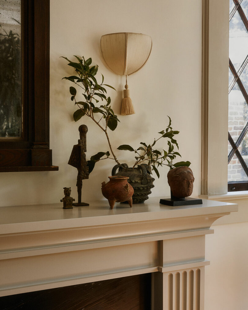 A Workstead sconce illuminates a mantelpiece adorned with a potted plant, small sculpture, carved figure, ceramic teapot, and a bust against a cream-colored wall.