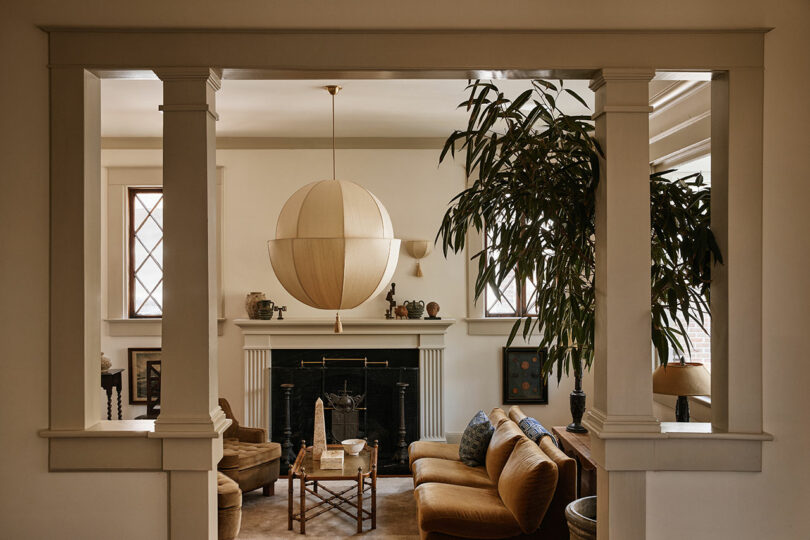 A cozy living room with tan sofas, a Workstead round paper lantern light, a fireplace, tall plant, and decorative objects on the mantel.