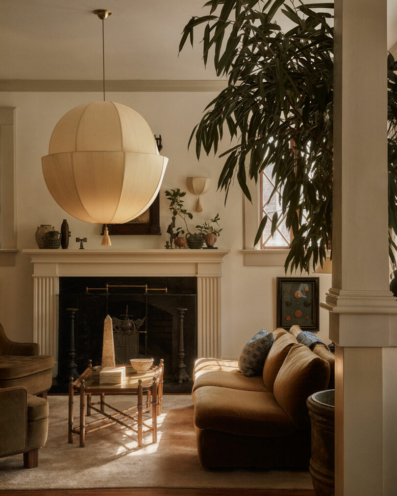 A warmly lit living room with a large Workstead globe-shaped pendant light, brown sofa, armchairs, fireplace, plants, and decor items on a mantelpiece.