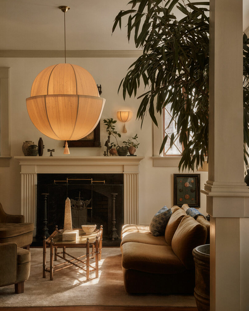 A warmly lit living room features a large Workstead spherical pendant lamp, a sofa, armchairs, a coffee table, and a fireplace with decorative objects. A tall indoor plant stands in the foreground.
