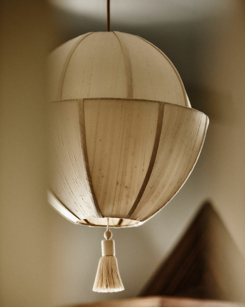 A beige, dome-shaped paper lantern by Workstead with vertical panels and a tassel hangs from the ceiling indoors.