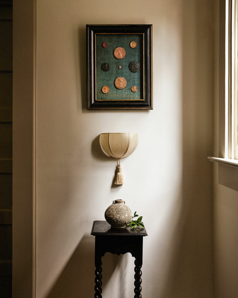 A small table with a round ceramic vase and greenery stands against a wall beneath a Workstead sconce and a framed display of coins, as natural light pours in from a nearby window.