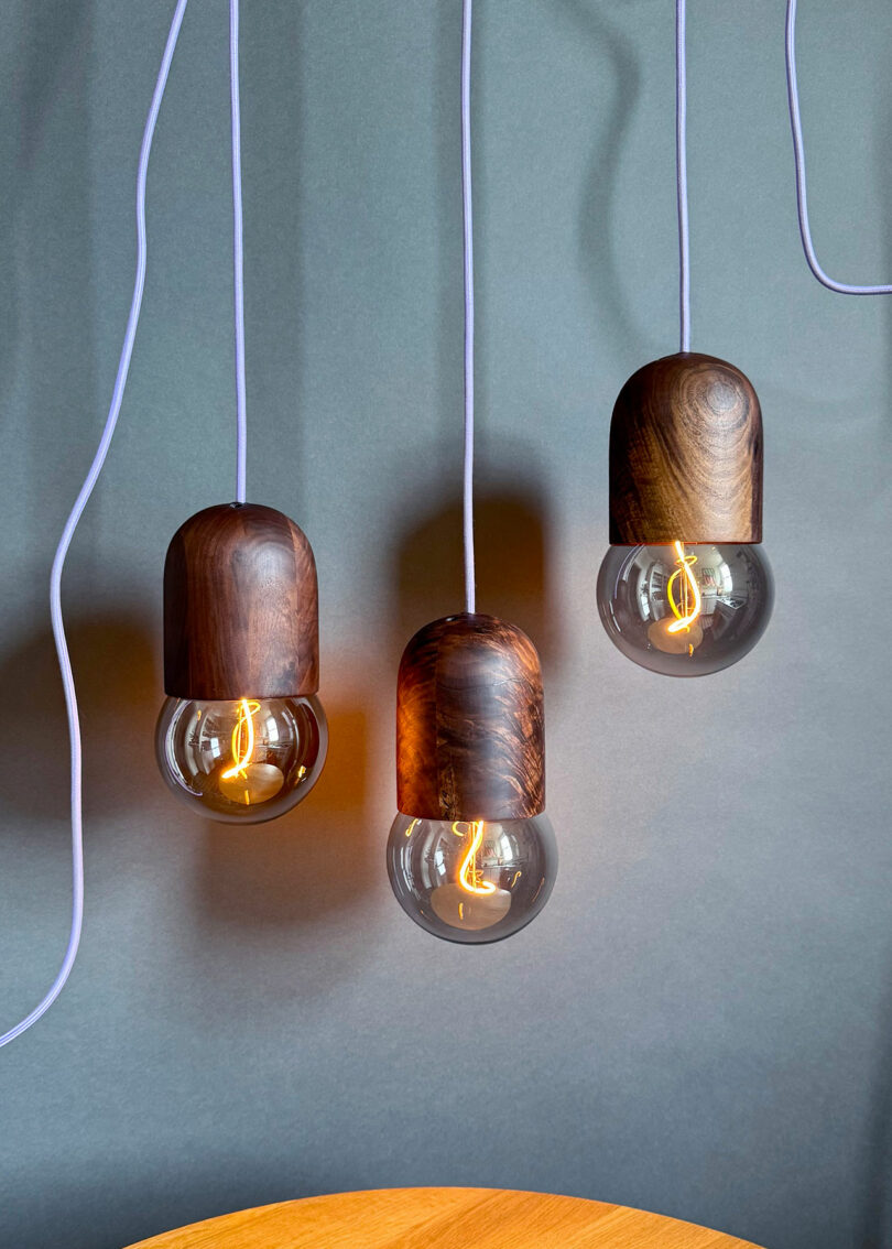 Three Light Bean pendant lights by Katerina Kopytina, featuring wooden tops and exposed filament bulbs, hang against a gray background with visible white cords.